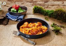 Изображение для онлайн-магазина активной жизни с одеждой, обувью для походов и путешествий, фрикадельки с макаронами Trek'n Eat на фоне гор и туристического снаряжения